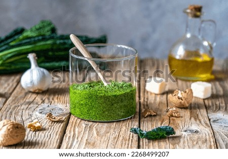 Homemade kale pesto served in a glass jar on wooden surface with grey background and basic ingredients - kale, walnuts, parmesan, garlic and olive oil