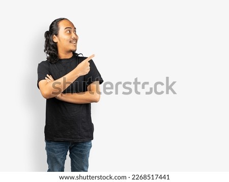 a young man with crossing left hand and pointing to the right with his right hand and smiling expression. A curly long hair guy with smile excited expression use black t-shirt and blue jeans standing