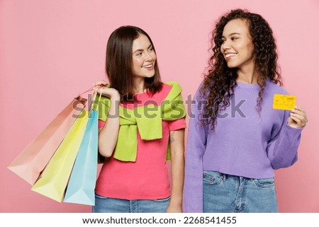 Young two friends happy women wears shirts hat hold in hand paper package bags after shopping together use credit bank card isolated on plain light pink background. Black Friday sale buy day concept
