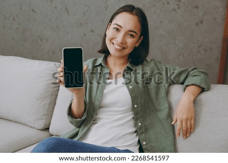 Young fun woman wears casual clothes hold use mobile cell phone with blank screen workspace area sits on grey sofa couch stay at home hotel flat rest relax spend free spare time in living room indoor