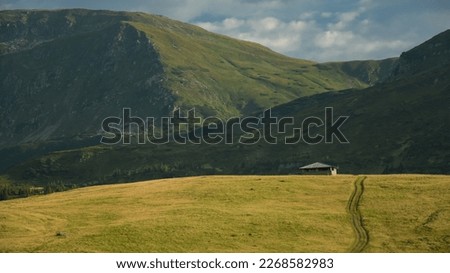 Sunset above the alpine pastures of Rodna Mountains. Rodna Peak is lighted by the sunlight. Traces of a car lead to a wooden cabin. Sunset time, Carpathia. Royalty-Free Stock Photo #2268582983