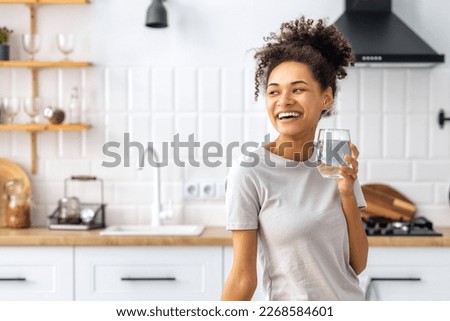Portrait of a beautiful African American young woman drinking water standing in the kitchen at home looking away and smiling, wellness healthy food concept Royalty-Free Stock Photo #2268584601