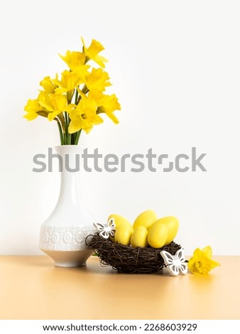 Yellow easter eggs in nest and bunch of daffodil flowers in white vase on beige table and white background. Happy Easter and holiday preparation. Festive decoration. Vertical orientation