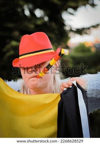 Portrait of a beautiful middle aged blonde caucasian woman with a cap on her head holding a Belgian flag and blowing a whistle celebrating independence day and winning a football match in the backyard