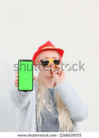 Blurred portrait of a caucasian blonde woman in a cap with a Belgian flag, sunglasses, a pipe and holding out her hand with a mobile phone with a green screen in the backyard of her house against the 