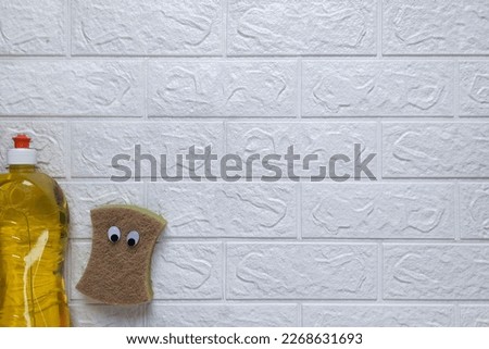 Kitchen washcloth with eyes and household chemicals. Top view. Copy space. Cartoon sponges for washing dishes with detergent on white brick wall background. Flat lay. Template for banner, advert.