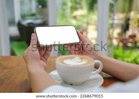 Mockup image of a woman holding and using mobile phone with blank desktop screen with coffee cup on the table in cafe