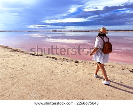 Latin adult woman with shorts, pink shirt, hat and sunglasses walks on the sand next to the pink colored lagoon with a high concentration of salt, Las Coloradas in Yucatan Mexico Royalty-Free Stock Photo #2268695845