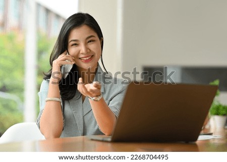 Cheerful asian female manager in stylish suit having phone conversation and smiling to camera.