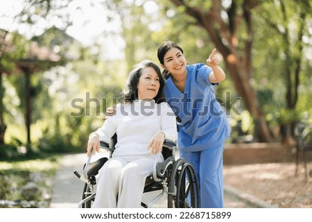 Elderly asian senior woman on wheelchair with Asian careful caregiver and encourage patient, walking in garden. with care from a caregiver and senior health insurance. in sun light

