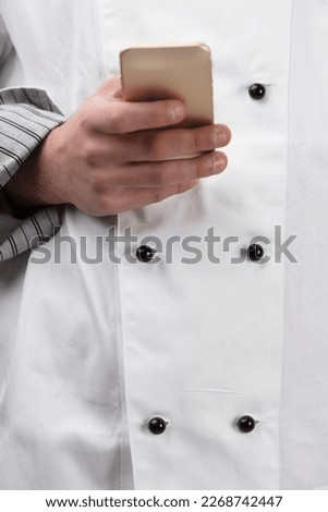 The hand of a professional cook or chef holds a smartphone. In the background is his official uniform, white with double parallel black buttons, a sign of experience and skill.