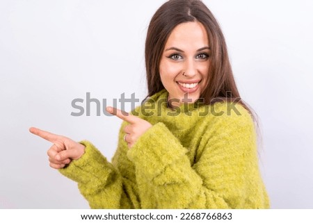 Attractive woman smiling pointing fingers at copy space on white background, green wool sweater