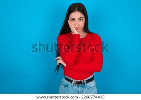 Very bored young brunette woman wearing red shirt over white studio background holding hand on cheek while support it with another crossed hand, looking tired and sick,