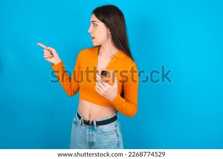 Stunned young woman wearing orange crop top over blue studio background points sideways right copy space, recommends product, sees astonishing thing
