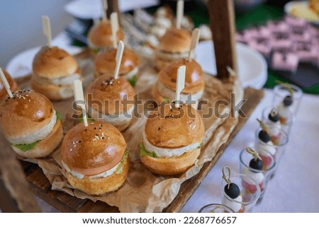 Abstract blurry food background. Small burgers with meat, salads cheese and tomatoes on wooden plate during summer barbecue party outdoors. Friends preparing food, chatting and having drinks together