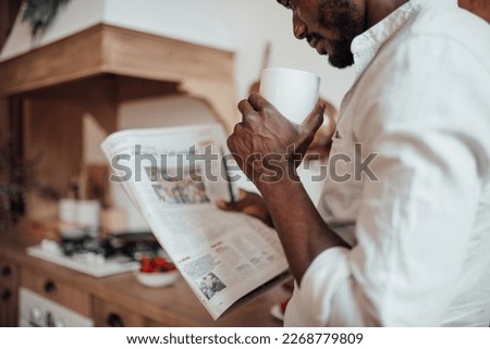 Candid millennial authentic diverse portrait of african american man relax time in neutrals tones kitchen. Brown skin male drink tea at breakfast domestic life and morning routine reading newspaper Royalty-Free Stock Photo #2268779809