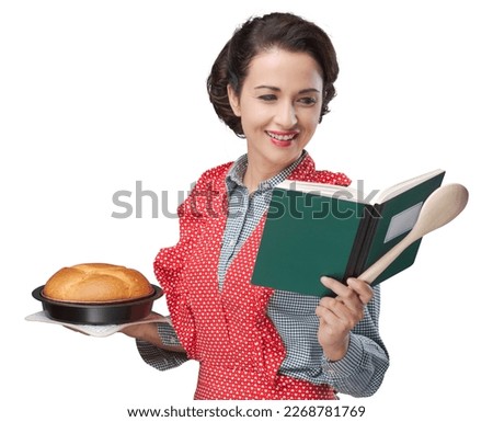 Smiling retro housewife holding a cookbook and a baking tin with a homemade cake Royalty-Free Stock Photo #2268781769