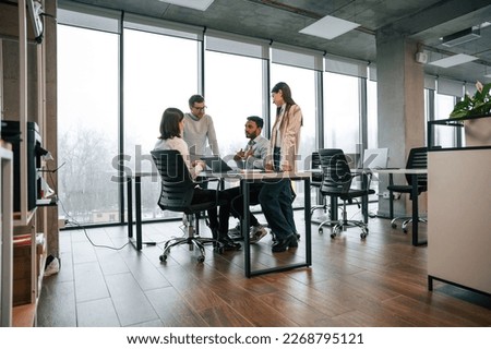 Against big windows by the table. Four people are working in the office together.