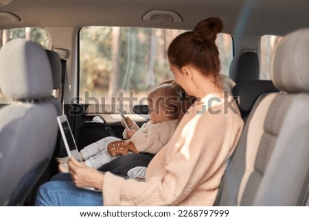 Image of Caucasian woman working on laptop while sitting with her baby daughter in safety chair on backseat of car, child watching cartoons on tablet, family traveling.