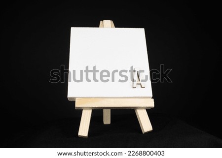 A wooden capital letter in bottom corner and white blank painting canvas resting on an miniature artists easel isolated on a black background