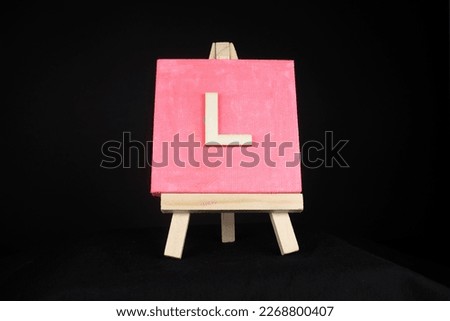 L wooden capital letter and pink blank painting canvas resting on a miniature artists easel isolated on a black background