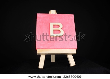 B wooden capital letter and pink blank painting canvas resting on a miniature artists easel isolated on a black background