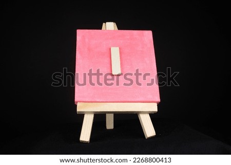 I wooden capital letter and pink blank painting canvas resting on a miniature artists easel isolated on a black background