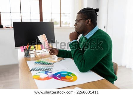 Thoughtful Black Male Designer Holding Color Palette Working Sitting Near Computer At Desk In Modern Office. Side View Of Man Thinking Choosing Color Scheme For Website. Professional Design Career