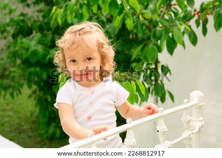 A child covered in strawberry juice stands on a white mettal bed in the garden