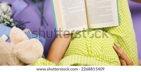 Beautiful pregnant woman dressed in a green dress is reading a book about motherhood, on a purple background in the studio. High quality photo