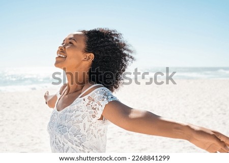 Healthy young african woman with curly hair standing at beach with hands outstretched and looking up. Happy young black woman with open arms feeling the sea breeze with copy space. Beautiful girl. Royalty-Free Stock Photo #2268841299
