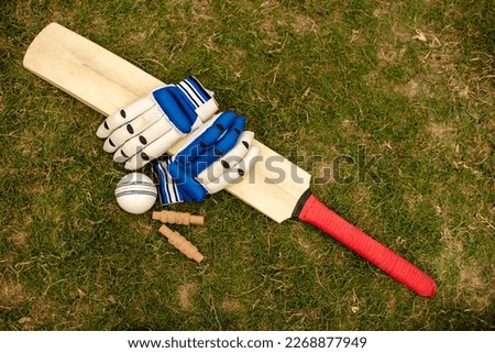 Cricket bat, ball gloves and bail are on green playing field