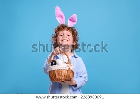 Portrait of happy boy in cute bunny ears headband holding wicker basket with Easter eggs on light blue background