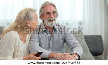 Caucasian elderly couple are sitting on the sofa and watching television in the living room. Senior grandmother is using a remote control to change channels for watching boxing show with her husband.