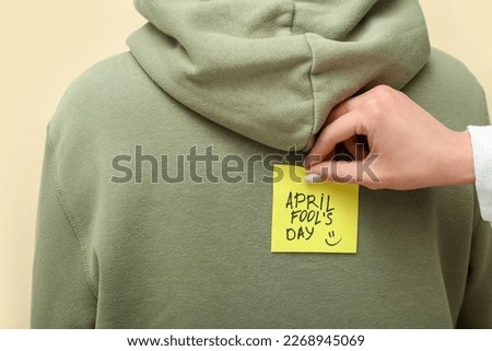 Woman sticking paper with text APRIL FOOL'S DAY on friend's back against color background, closeup Royalty-Free Stock Photo #2268945069