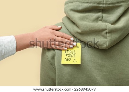 Woman sticking paper with text APRIL FOOL'S DAY on friend's back against color background, closeup Royalty-Free Stock Photo #2268945079