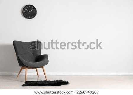 Stylish grey armchair, clock and black fur rug near white wall