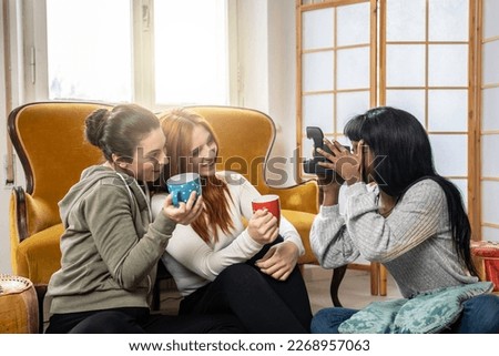 Cheerful multicultural female friends enjoying free time together at home - Happy young women while drinking coffee or tea and making photos with vintage camera in living room