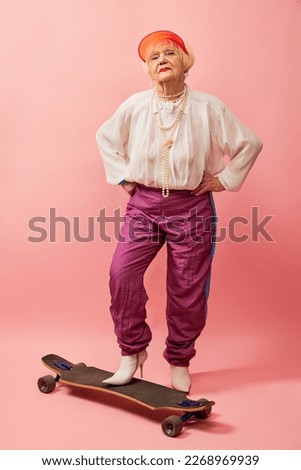 Beautiful old woman, grandmother in stylish clothes posing with skateboard over pink studio background. Concept of age, fashion, lifestyle, emotions, facial expression Royalty-Free Stock Photo #2268969939