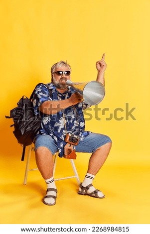 Portrait of fat mature man posing, sitting and shouting in megaphone over bright yellow studio background. Information. Concept of american style, culture, emotions, facial expression, travel