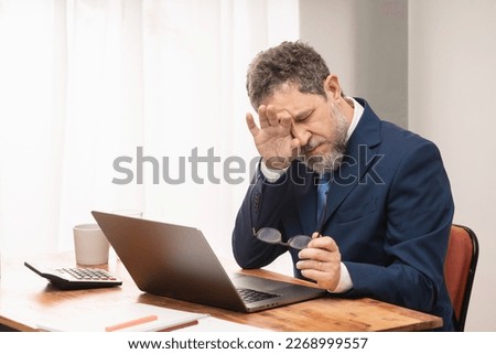 A tired middle-aged businessman in a blue suit and tie sits at his desk with his glasses in one hand, rubbing his eyes with the other. Prolonged computer use and age-related vision problems. Royalty-Free Stock Photo #2268999557