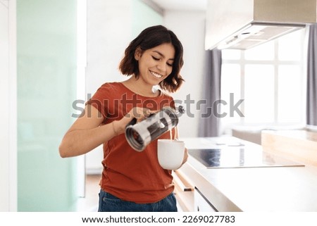 Portrait Of Young Smiling Arab Woman Making Morning Coffee At Home, Happy Millennial Middle Eastern Female Standing In Kitchen Interior, Holding French Press And Pouring Caffeine Drink To Cup Royalty-Free Stock Photo #2269027783