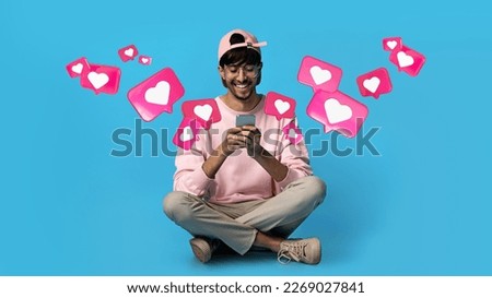 Glad young arabic guy in glasses has romantic chat with hearts on smartphone, enjoys message, sits on floor on blue studio background. Relationships and love remotely, app for dating, social networks Royalty-Free Stock Photo #2269027841