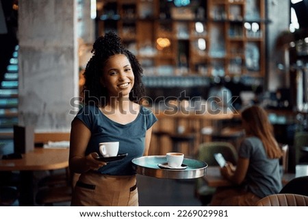 Happy African American waitress serving coffee while working in cafe ad looking at camera. Royalty-Free Stock Photo #2269029981