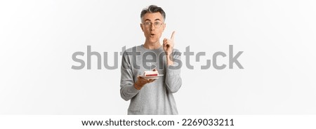 Image of happy middle-aged man, celebrating birthday, have great idea what to wish while blowing candle on b-day cake, standing over white background.