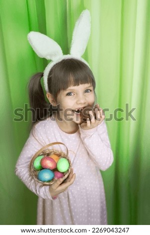 A portrait funny little girl with brown hair and a toothless smile, wearing bunny ears, eating chocolate egg and holding basket of Easter eggs in her hands. Copy space. . High quality photo