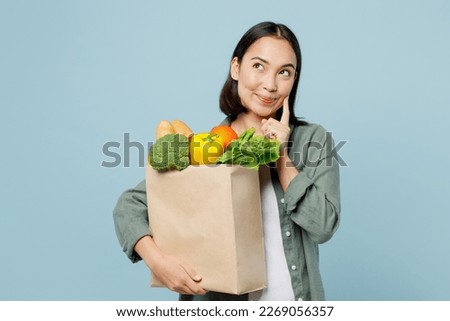 Young woman wear casual clothes hold brown paper bag with food products put hand prop up on chin look aside isolated on plain blue background studio portrait. Delivery service from shop or restaurant