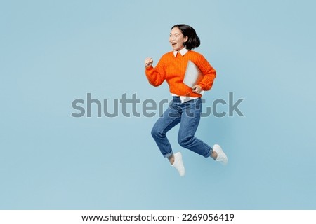 Full body side view excited young IT woman of Asian ethnicity wear orange sweater glasses jump high hold closed laptop pc computer run fast isolated on plain pastel light blue cyan background studio Royalty-Free Stock Photo #2269056419