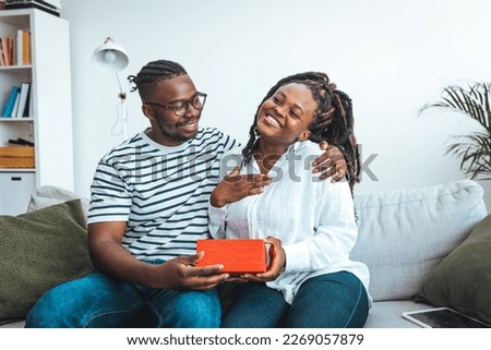 Shot of a loving husband giving his wife a gift. Boyfriend surprise his beautiful girlfriend with present while she is sitting on the sofa in the living room at home. Romantic couple with present.