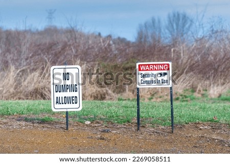 Horizontal shot of a No Dumping Allowed sign and a Warning Security Cameras In Use sign in a field.